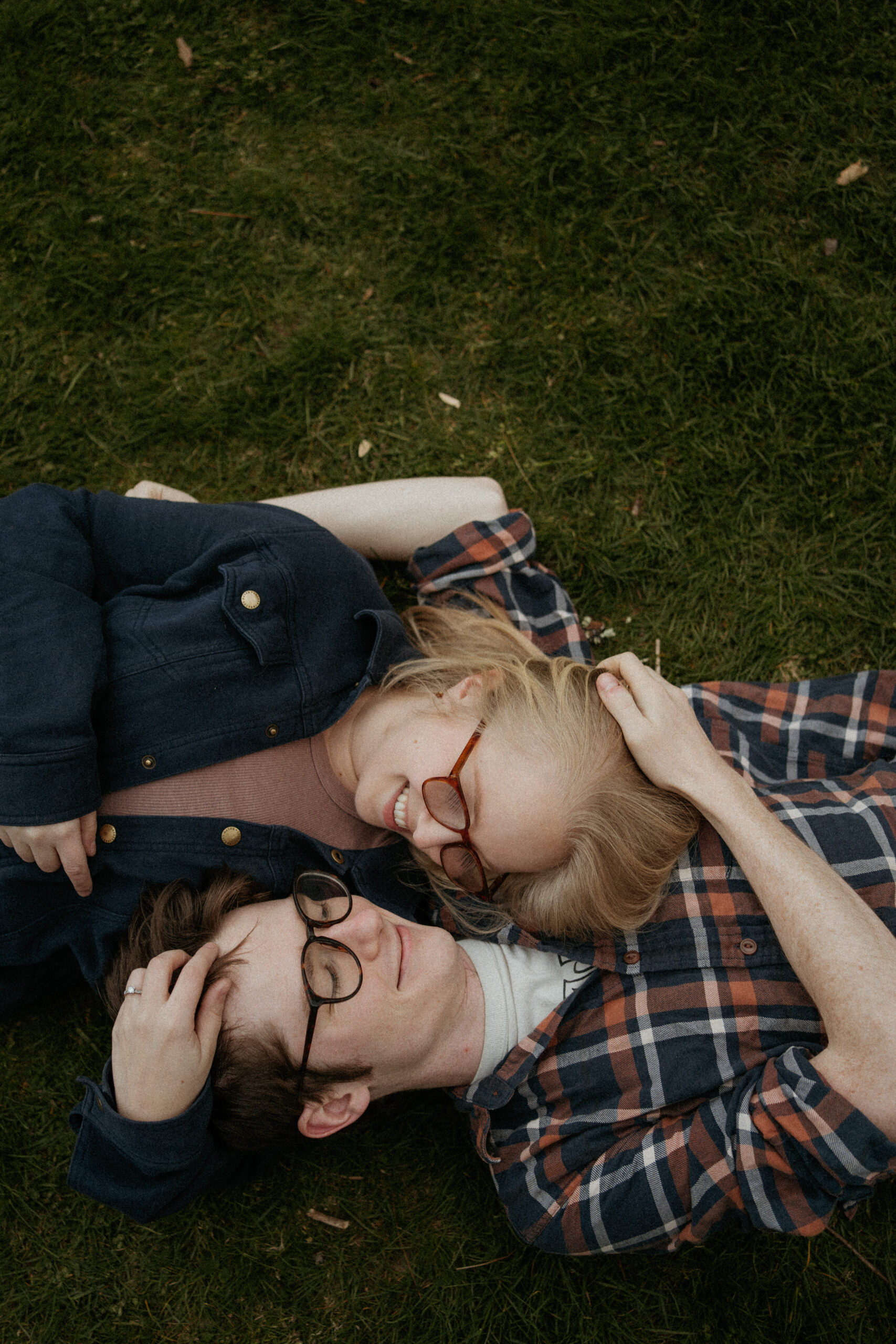 Engaged couple lies in the grass, heads resting on each other's shoulders, sharing tender gazes and gentle caresses during their heartfelt engagement photo session at Borderland State Park in Easton, MA.