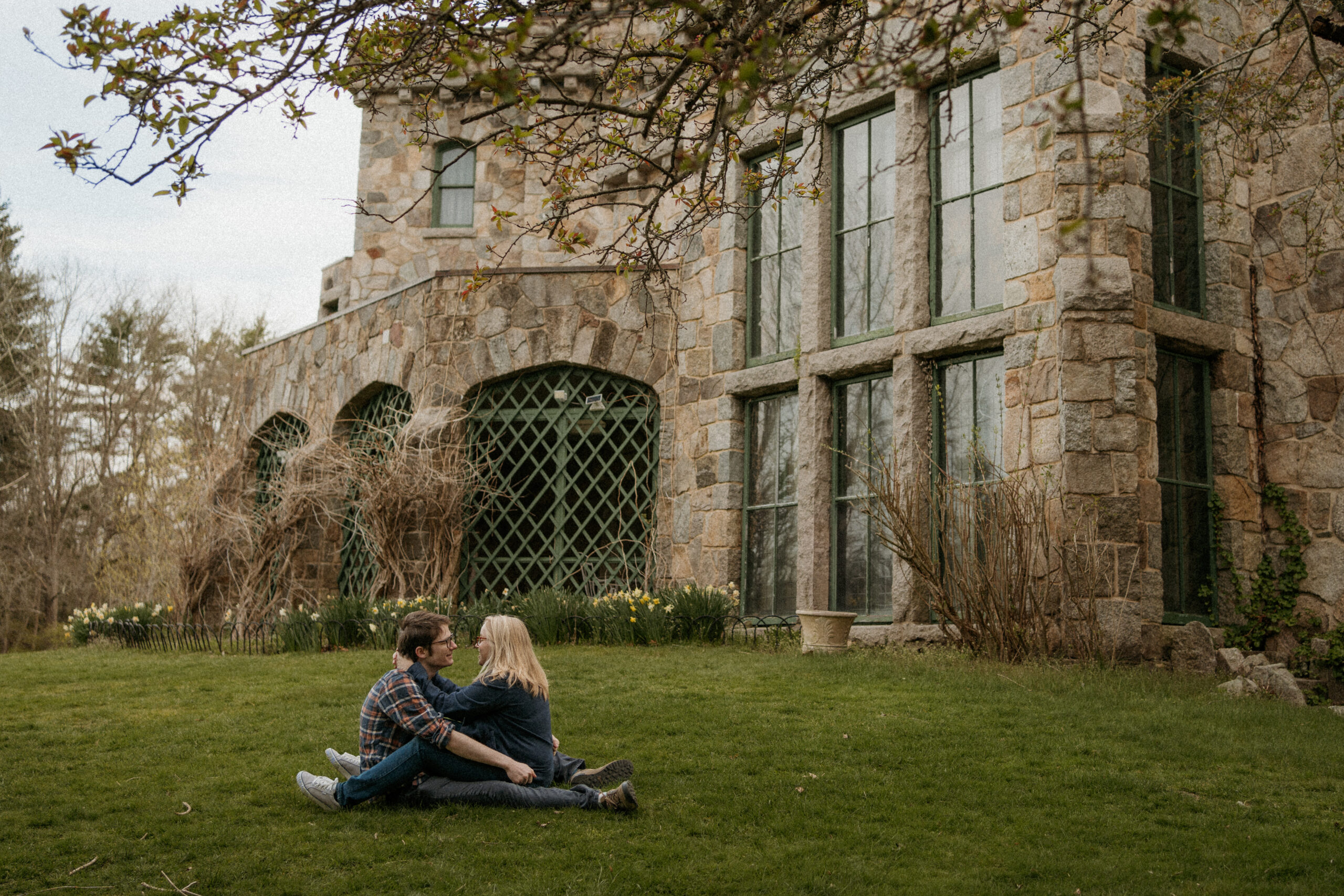 Engaged couple sits on the grass, facing each other, sharing a sweet hug in front of the charming Ames Mansion during their engagement session at Borderland State Park in Easton, MA.