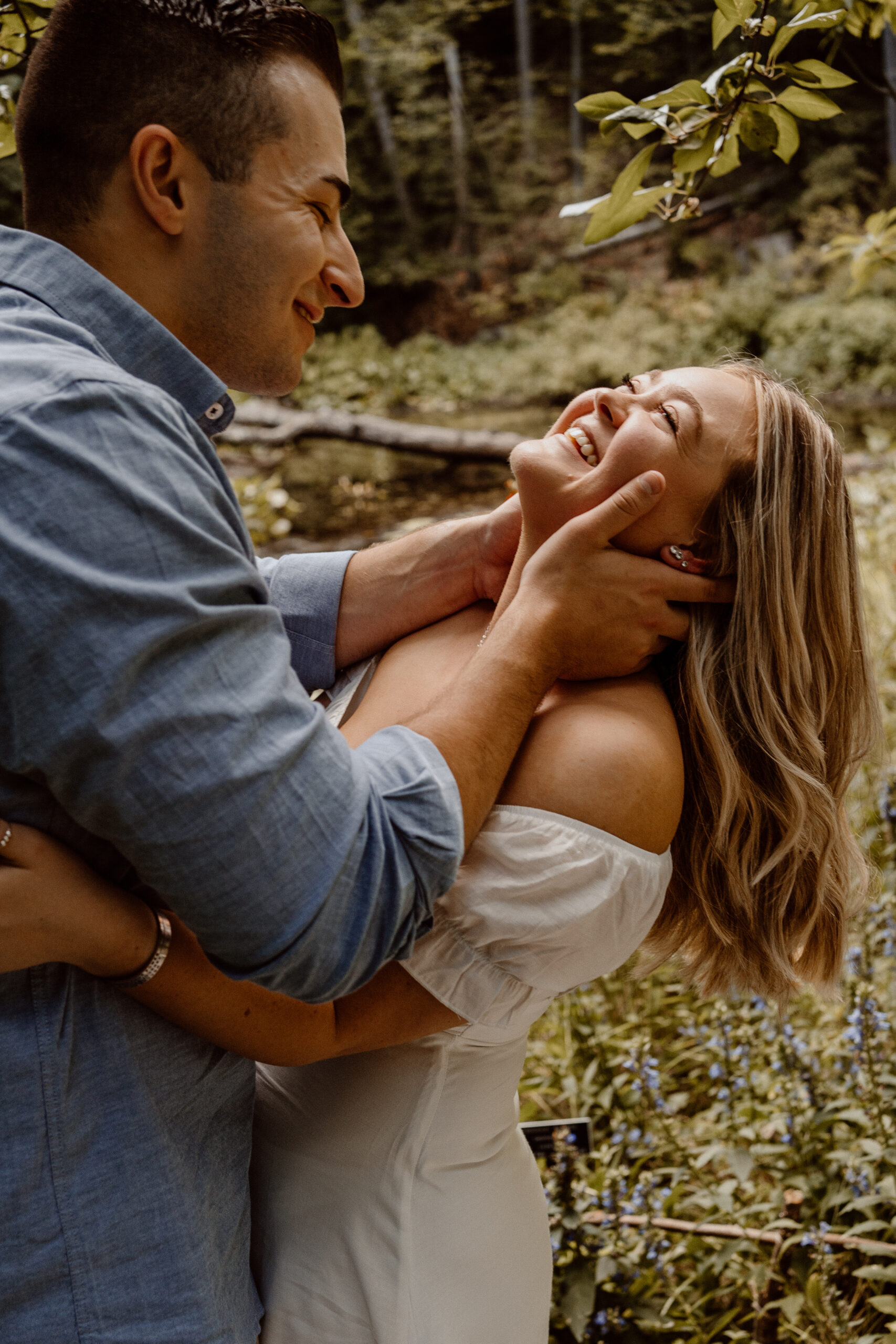 Happy engaged couple embraces and shares laughter amidst the scenic beauty of Garden in the Woods, Framingham, MA, surrounded by lush trees, capturing their love in a natural setting.