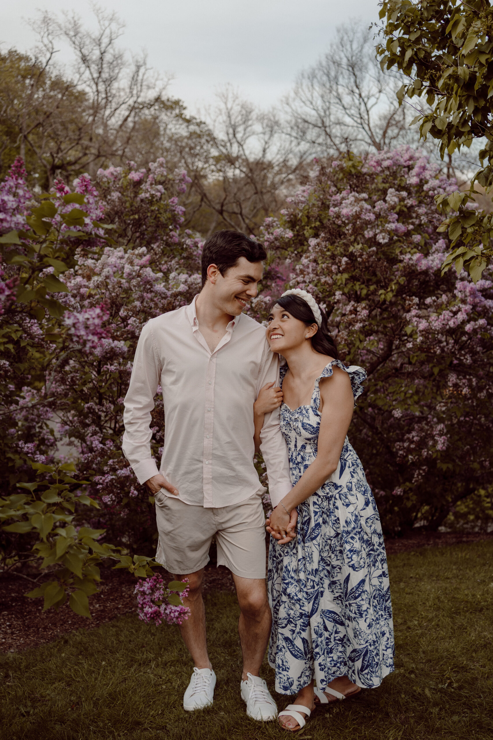 Engaged couple strolls hand in hand, sharing tender gazes amid the vibrant lilac trees during their delightful spring engagement session at the Arnold Arboretum in Boston, MA