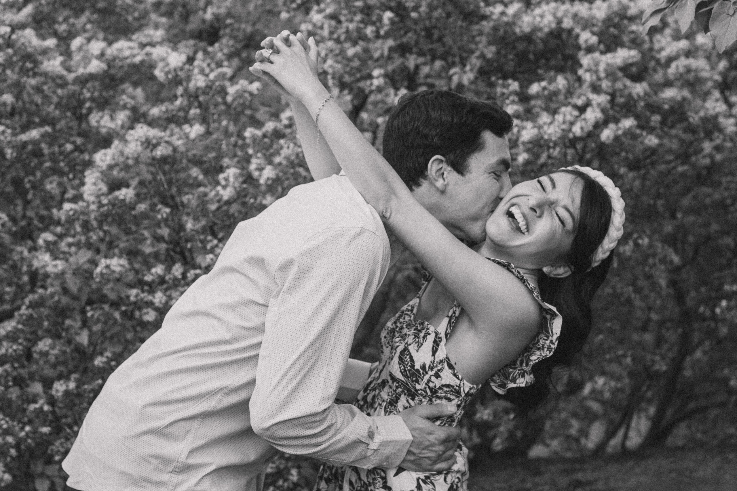 Engaged couple shares a joyful embrace, with the fiancé kissing her cheek as she laughs, surrounded by a field of lilac trees during their engagement session at Arnold Arboretum in Boston, MA