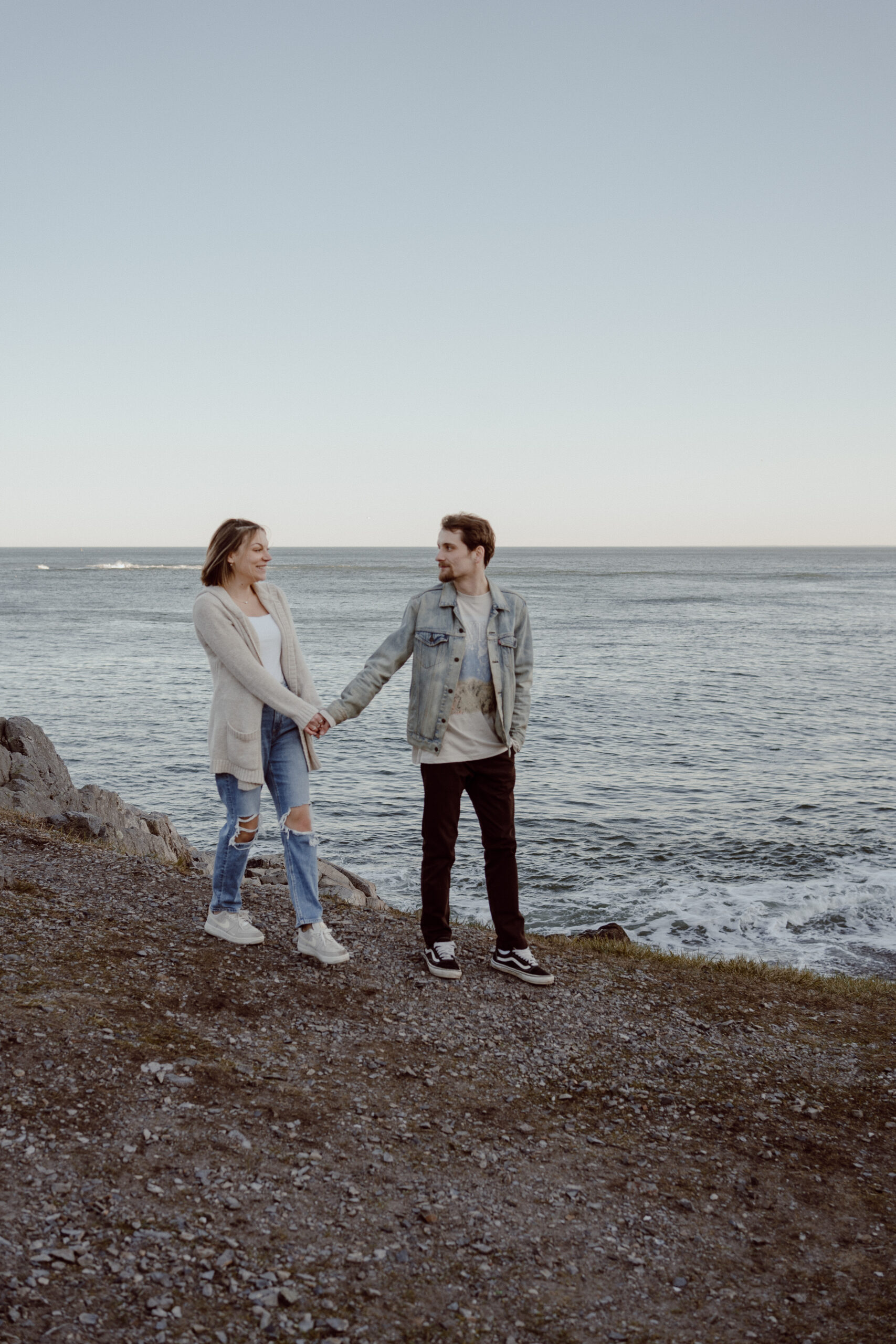 Engaged couple walks along the cliff's edge, sharing loving gazes during their romantic engagement session at Castle Rock, Marblehead, MA, capturing the breathtaking scenery and their affectionate bond