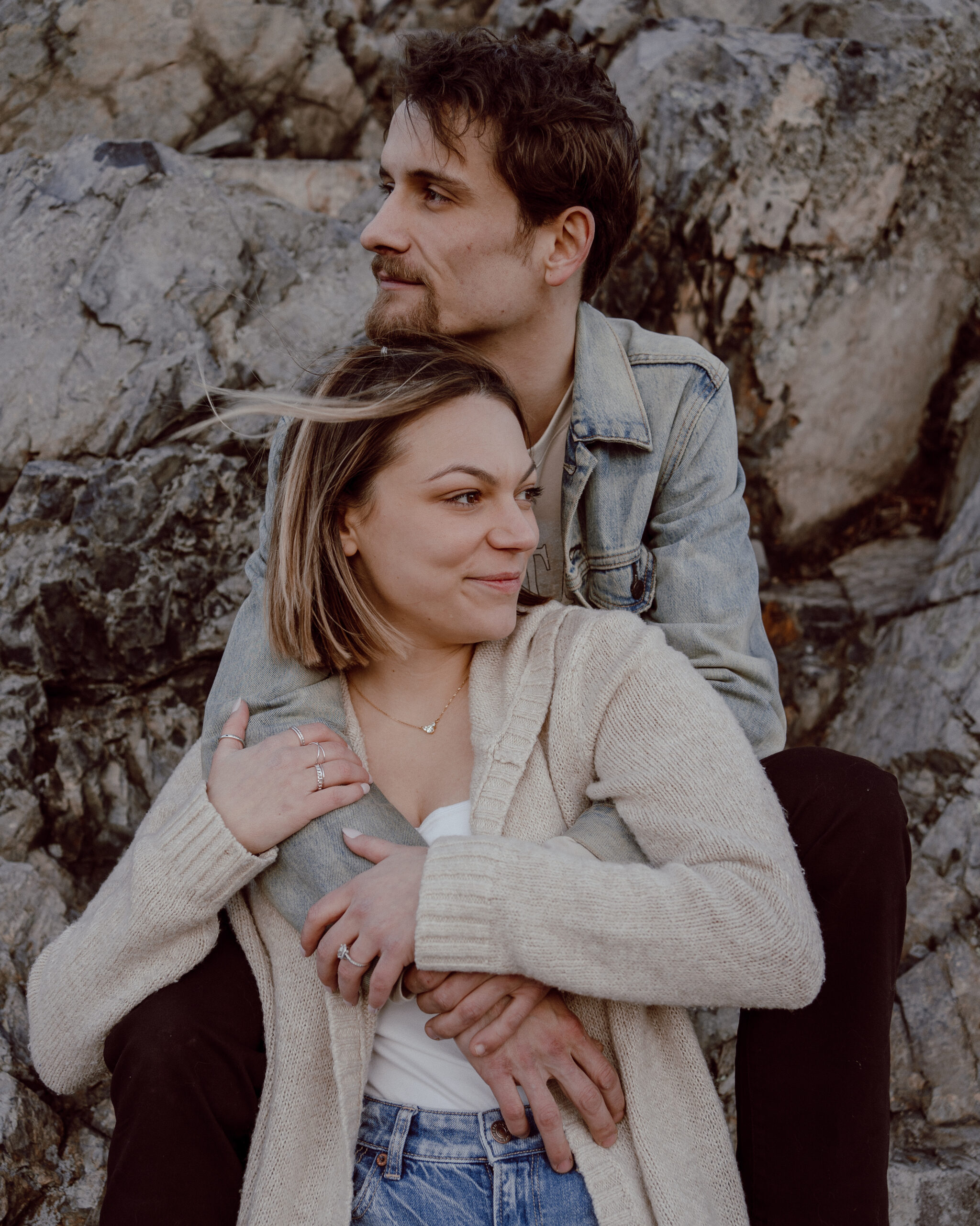 Engaged couple embraces against a rock backdrop, with the woman hugged from behind by her fiancé as they gaze in opposite directions during their engagement photo session at Castle Rock, Marblehead, MA.