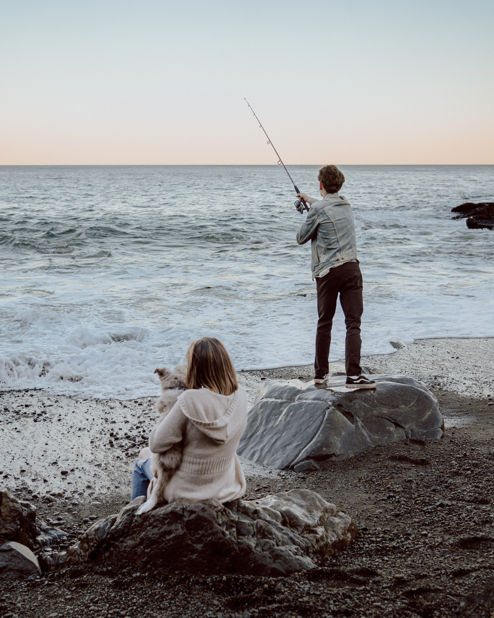 Engagement photo session at Castle Rock, Marblehead, MA: Fiancé fishes in the ocean while fiancée and their dog enjoy watching, capturing a relaxed moment by the coast.