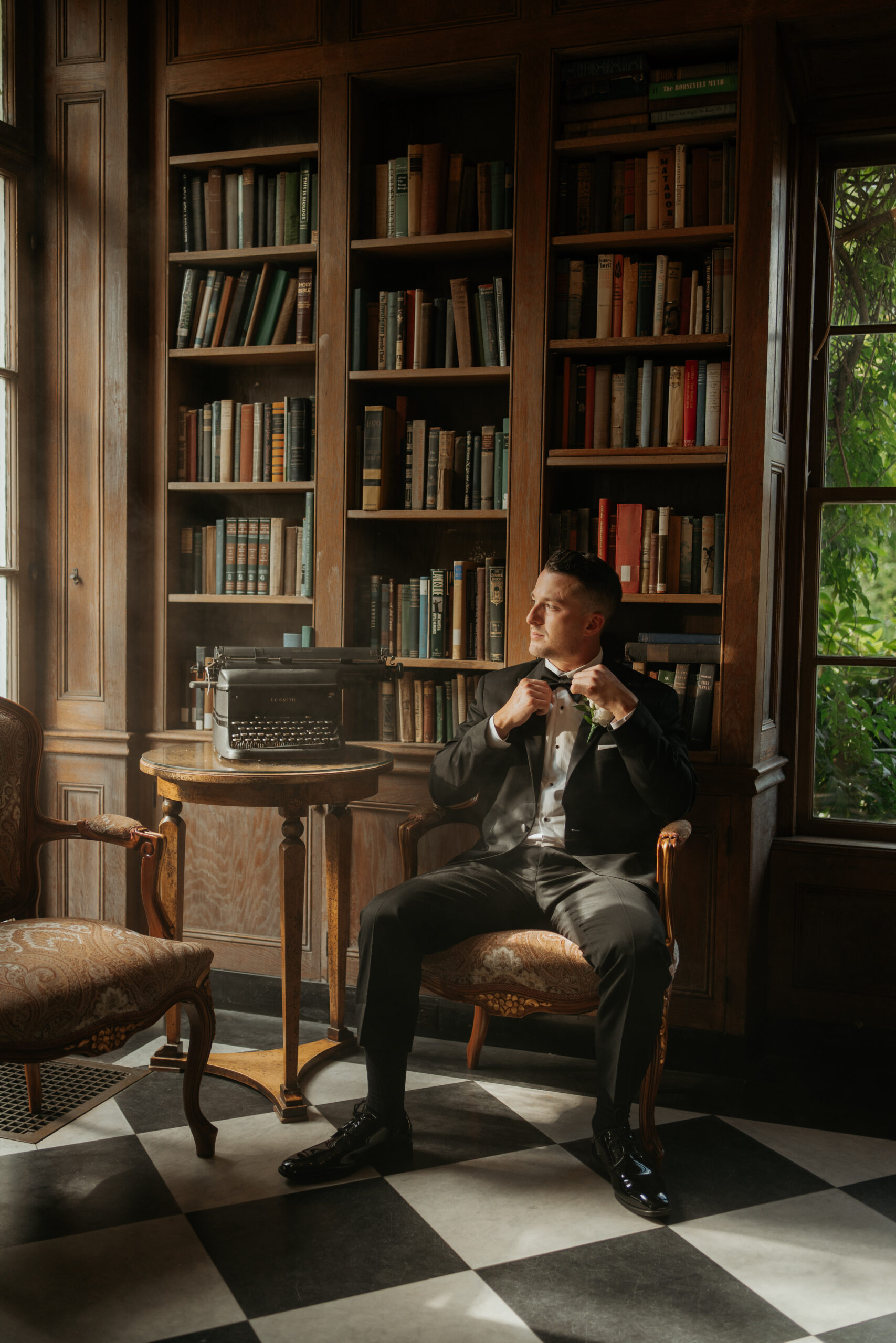 Groom sits and stares out the window as he adjusts his bowtie surrounded by haze in the study of Misselwood in Beverly, MA.