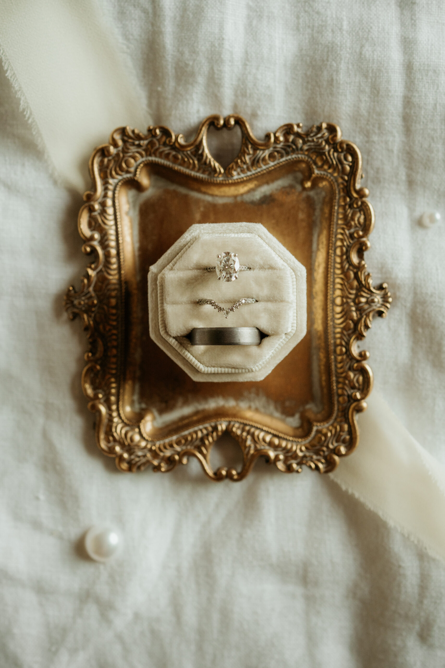 Elegant flat lay with oval engagement ring, tapered wedding ring & platinum wedding ring in a cream ring box set in a gold ornate rectangle ring dish. 
