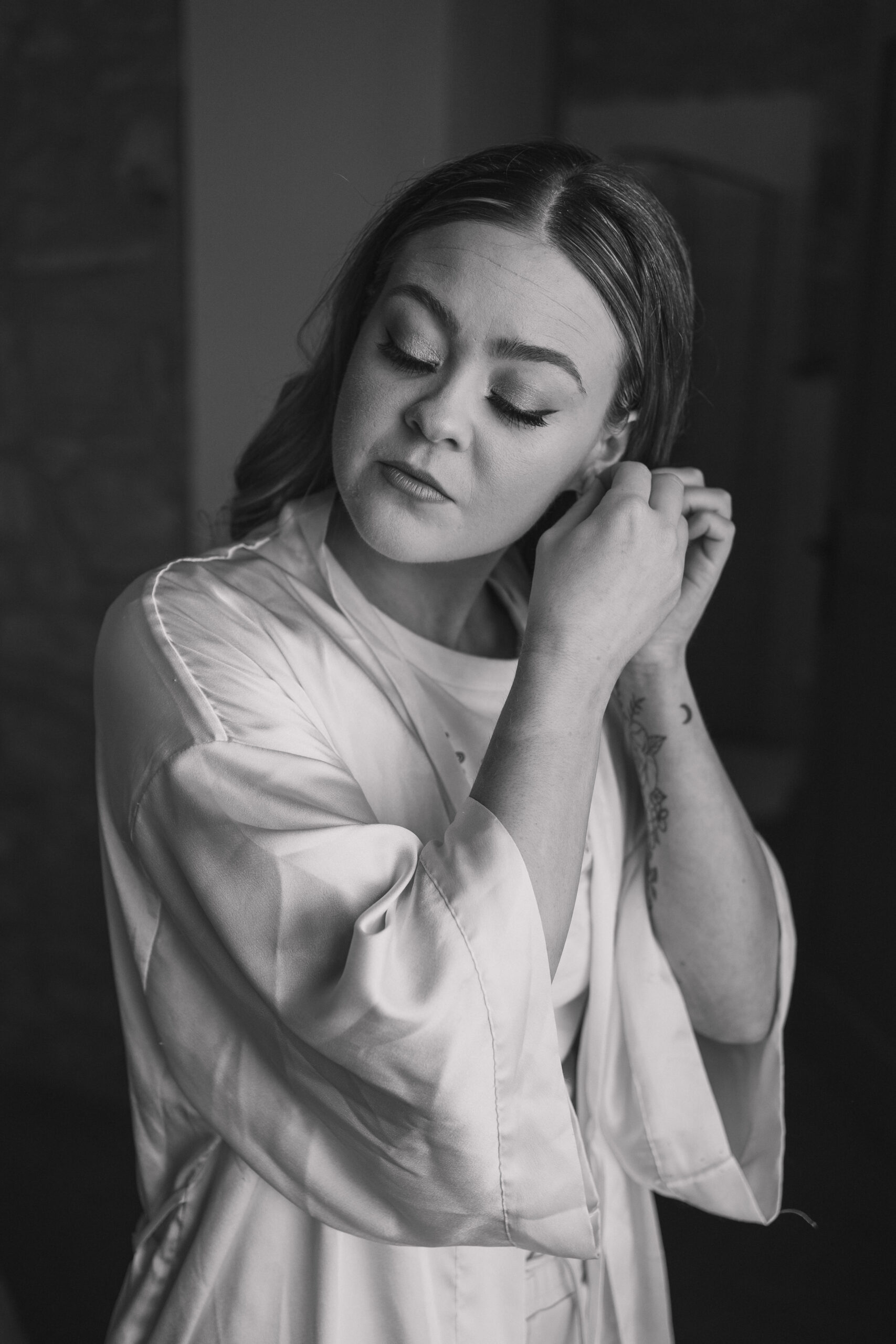 Bride in her white silk robe and pjs with her hair and makeup done, looks down as she puts her earring in 