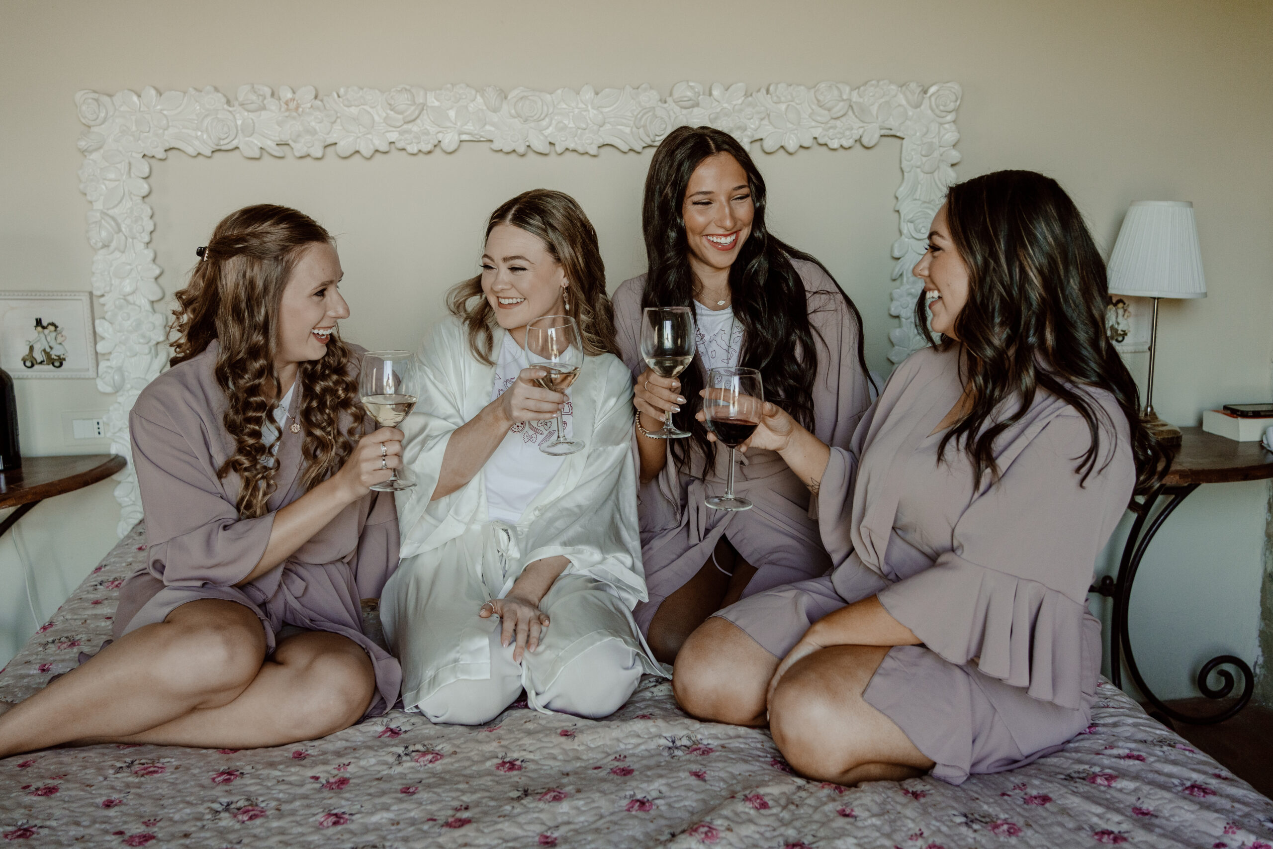 Bride is in her white silk robe and pjs on the bed surrounded by her three bridesmaids wearing mauve chiffon robes and pjs. They are laughing and looking at each other while cheersing their glasses on wine