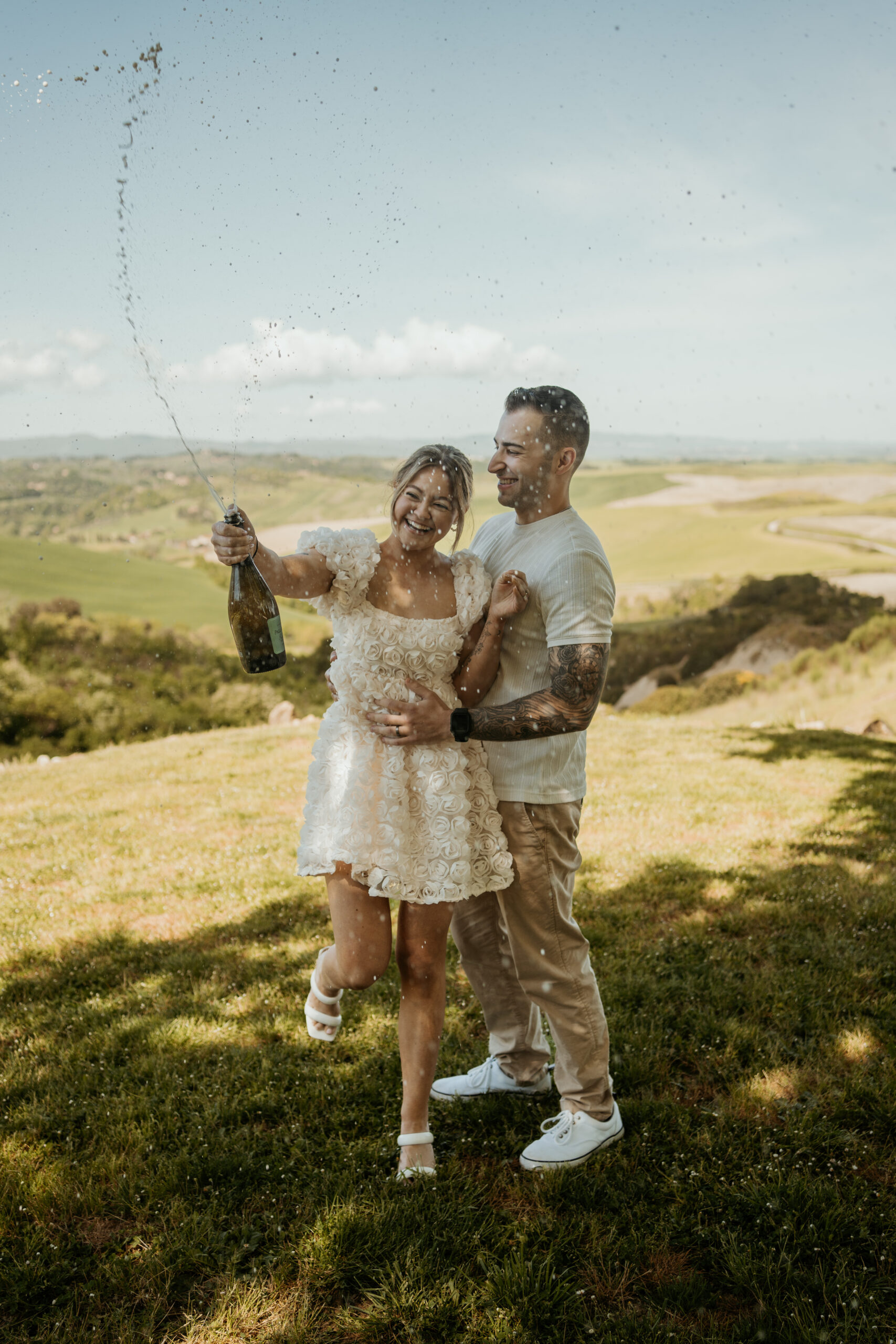 Bride and groom pop champagne and spray it out of excitement and celebration in anticipation of their Tuscan destination wedding day to begin. They are met with Tuscan rolling hills as their backdrop.
