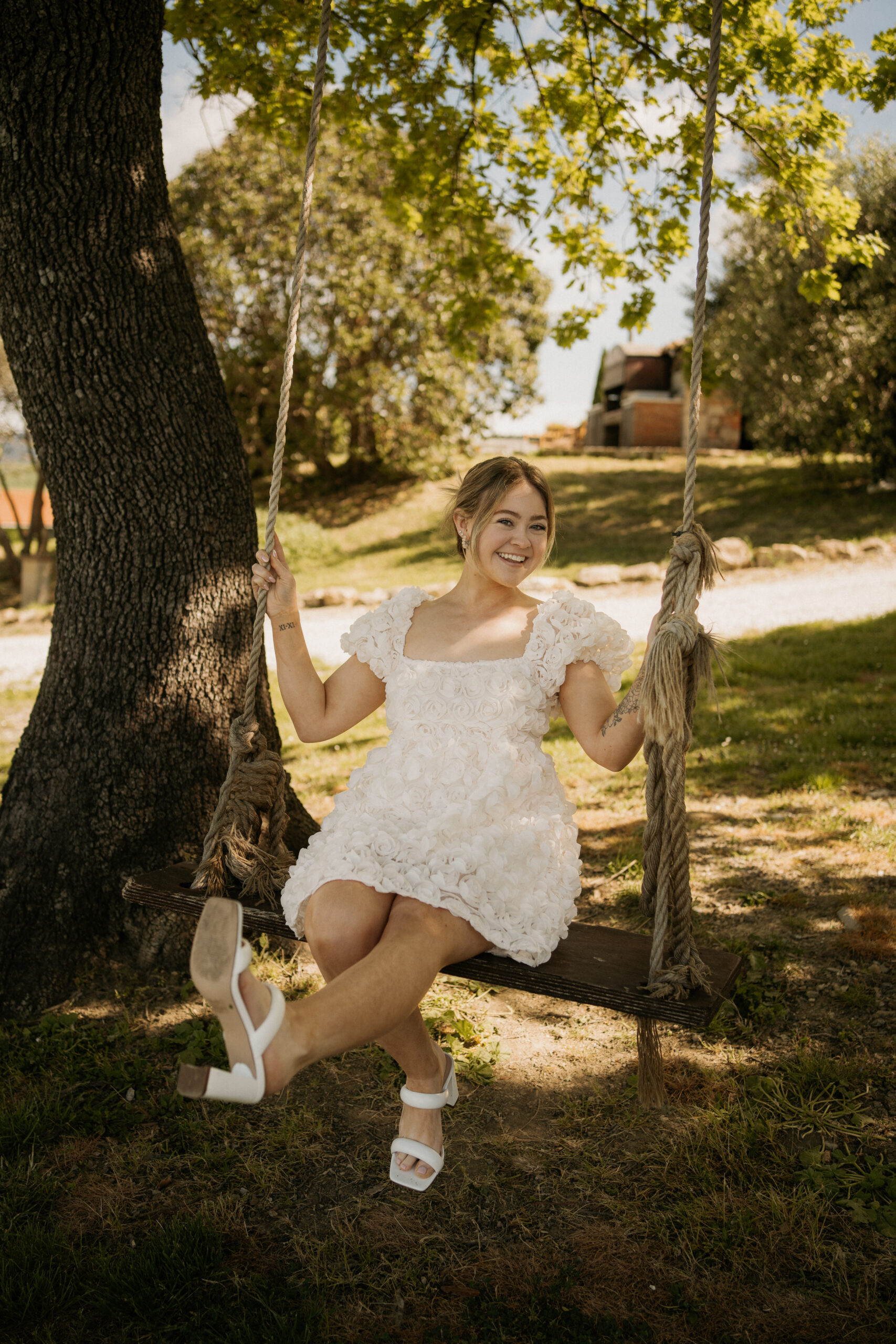 Bride, wearing a white rose bud cocktail dress, swings on a tree swing outside her Italian Villa Wedding Venue in Volterra