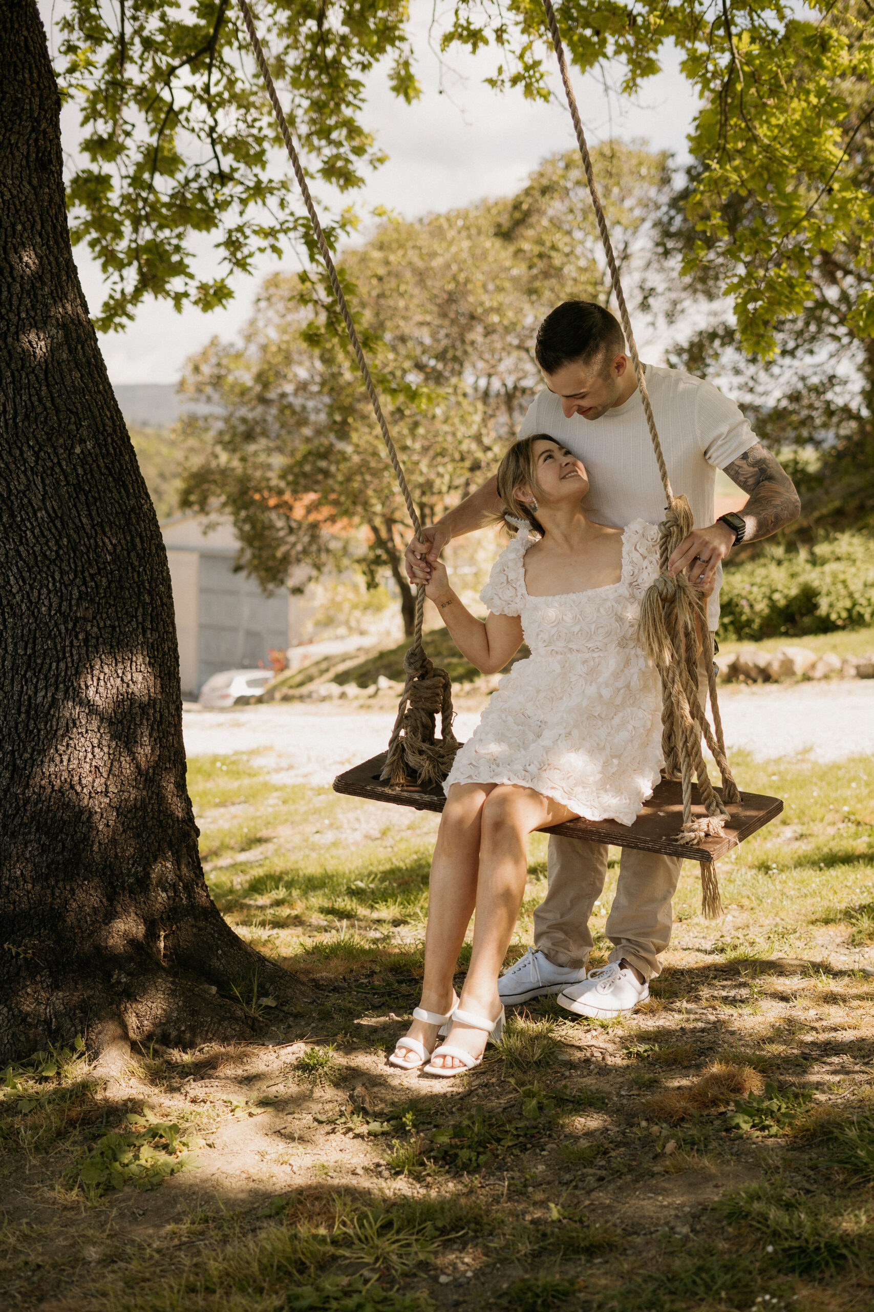 Bride, wearing a white rose bud cocktail dress, sitting on a tree swing  while the groom holds the rungs and looks down at her outside their Italian Villa Wedding Venue in Volterra