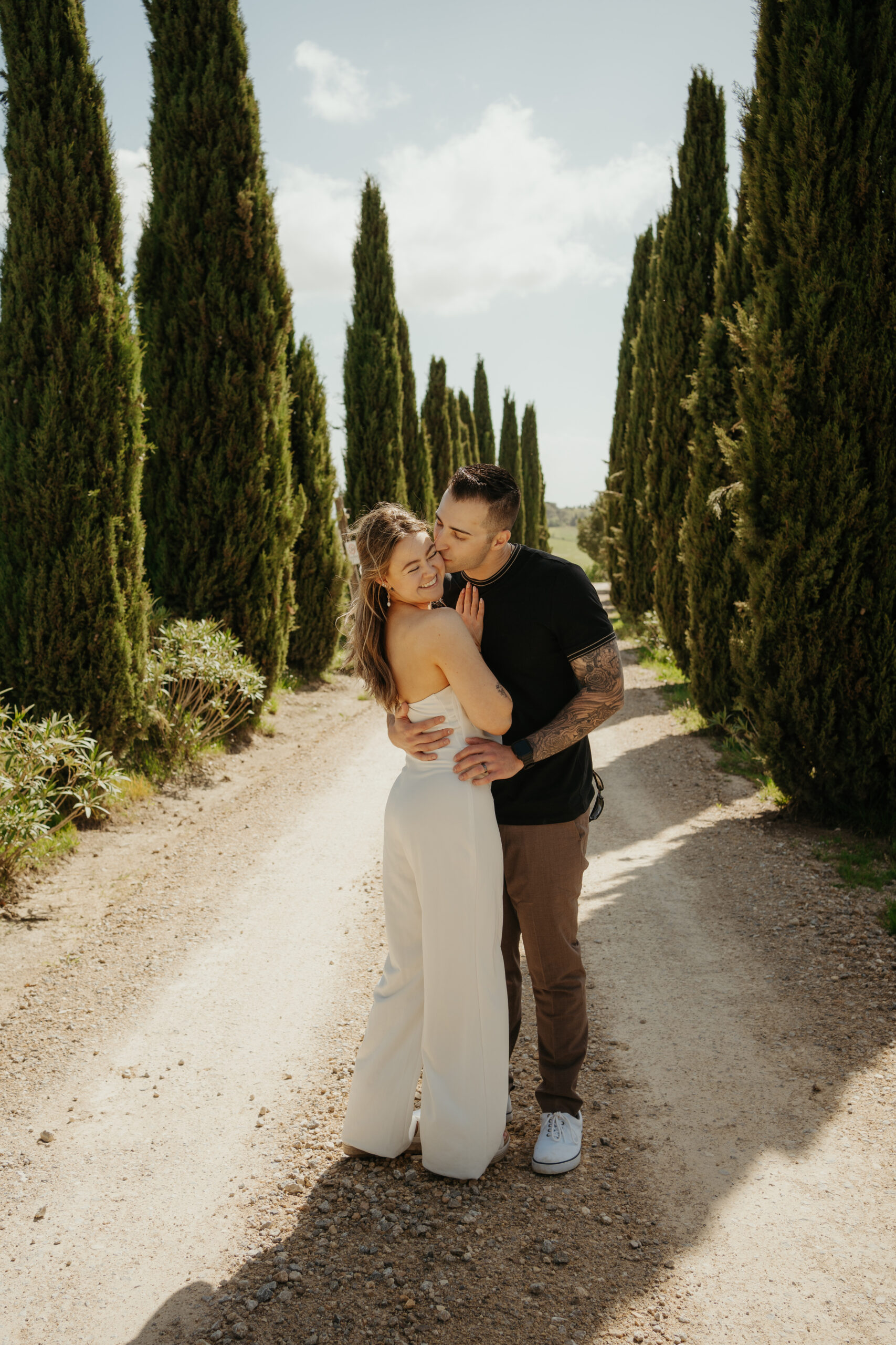 Bride and groom hug while groom kisses bride on the cheek in the middle of rows of Tuscan Pines during their Italy destination wedding excursion