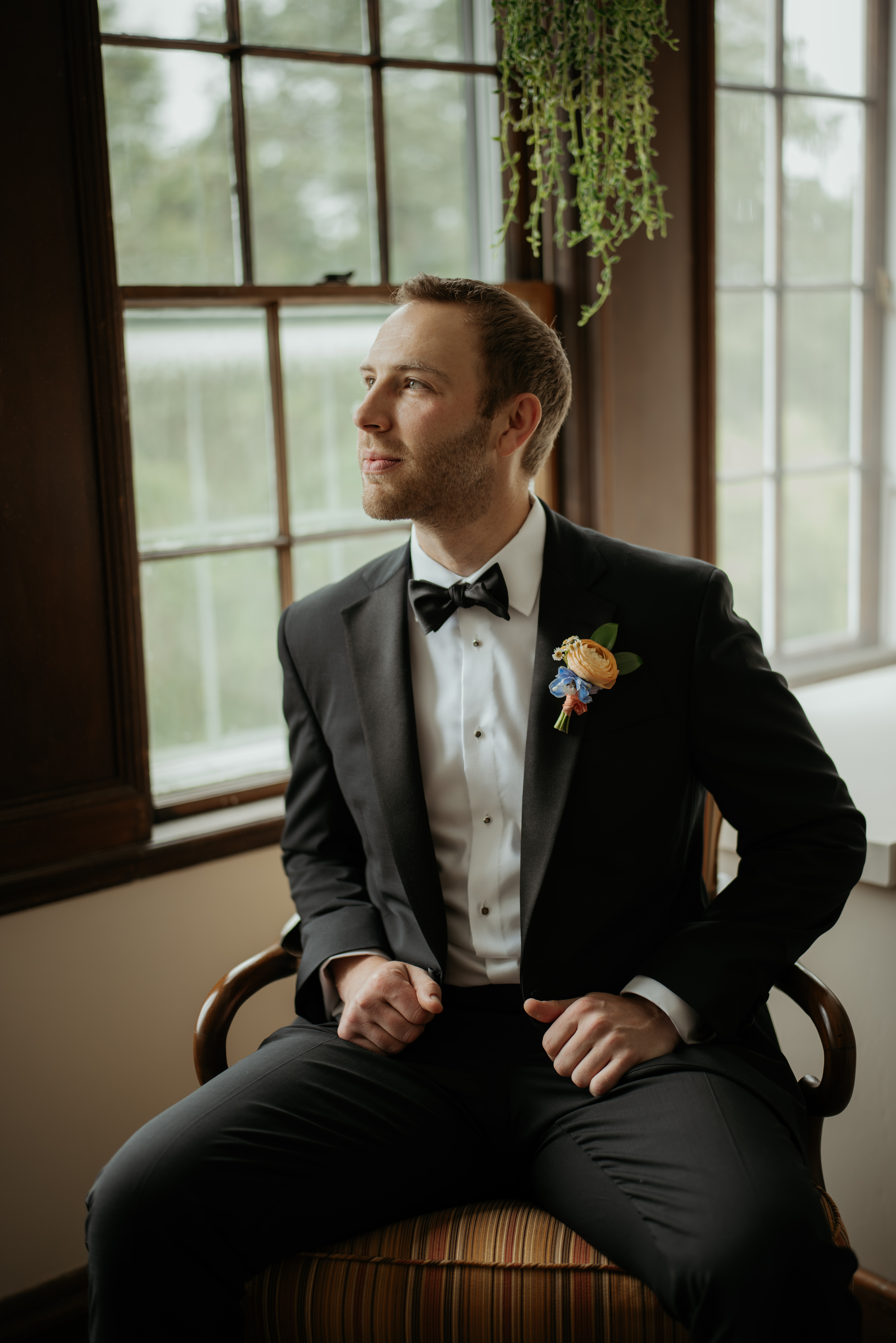 Groom dressed in his tux & bow tie sitting down in a chair, looking out the window. He looks excited for his summer garden party wedding day that is about to start.