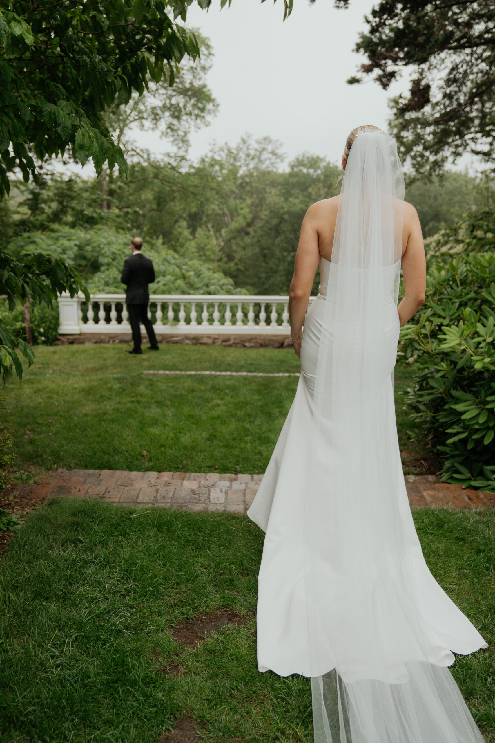 Bride approaches groom for their first look. Groom is standing with his back to the bride in the background and the bride is in the foreground. They are having their first look along the terrace of Long Hill in Beverly, MA.