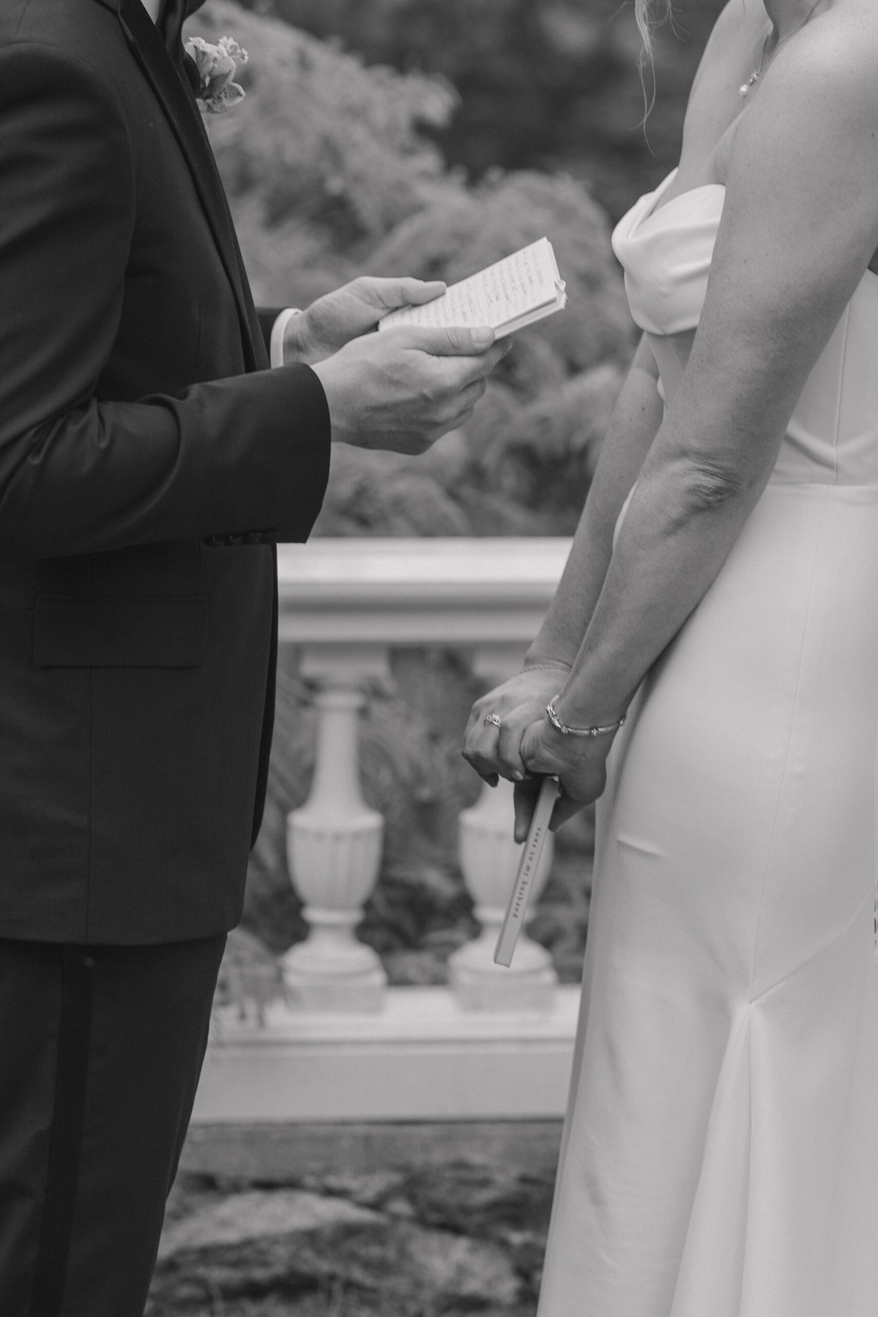 Hands of bride and groom. Groom has his vows open as he reads them privately and the bride has hers clasped in her hands down by her waist/