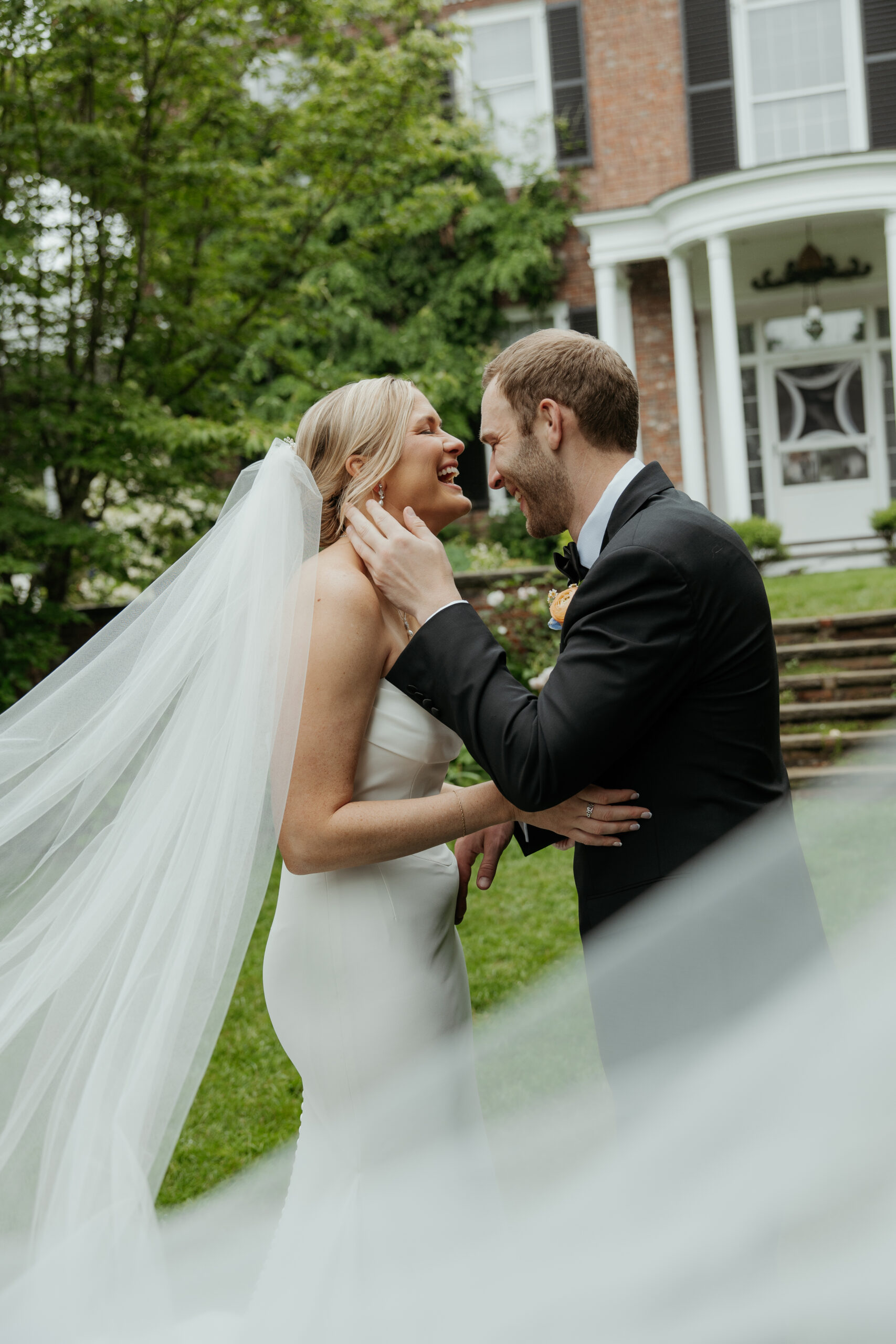 Bride and groom laughing together at their New England summer garden party wedding as the bride's veil swoops in front of the camera.