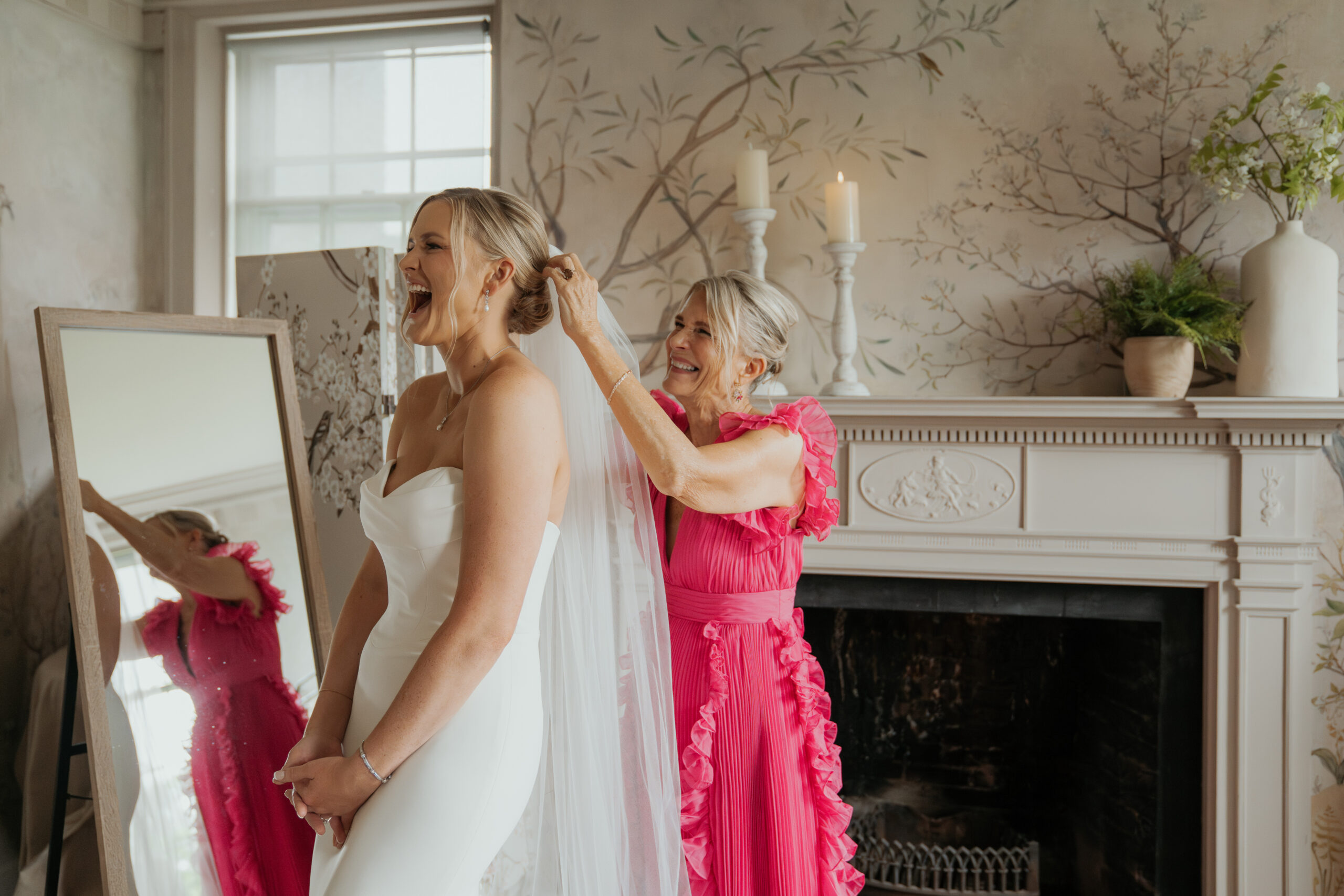 Bride's mother, dressed in a bright pink gown, puts bride's veil into brides hair. The bride is looking super happy with her mouth agape in a big smile. 
