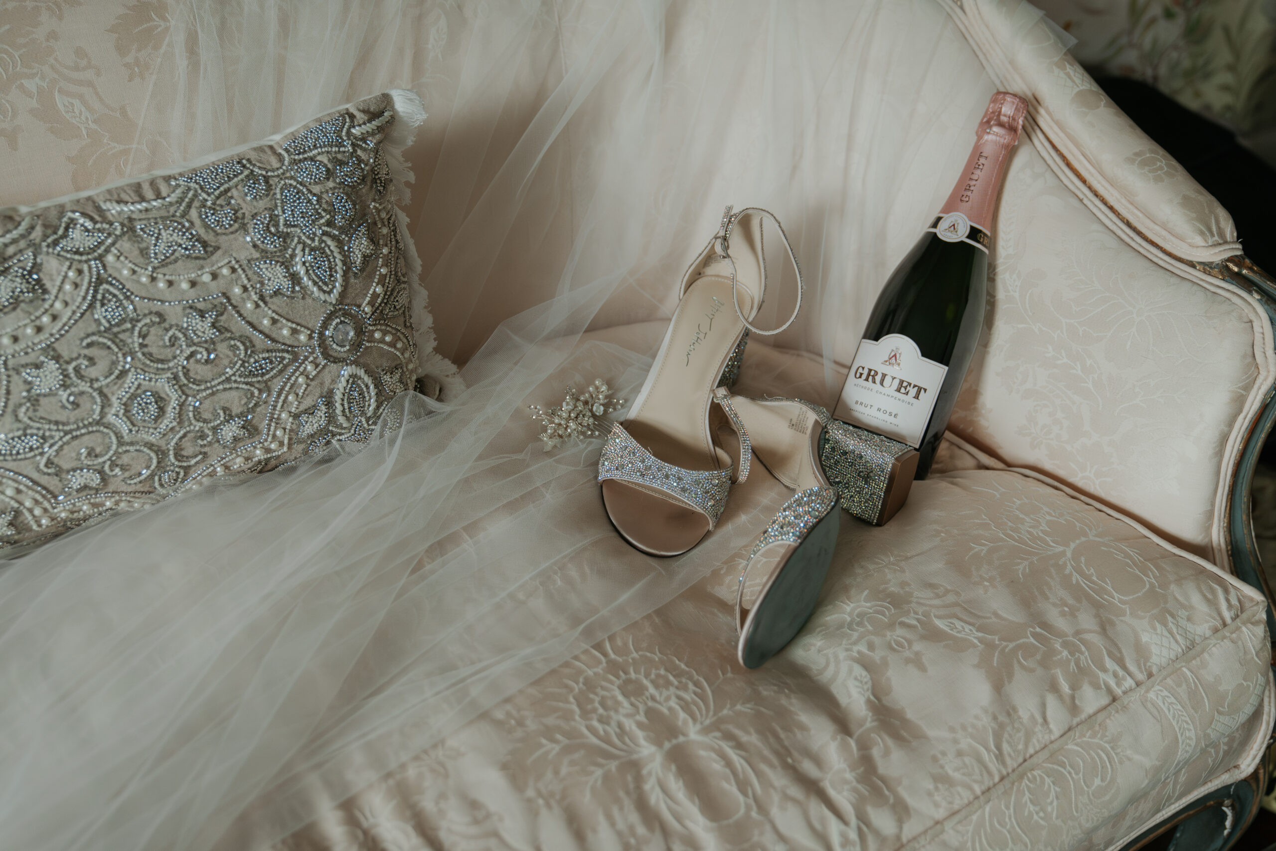 A bridal detail photo on a lavish white couch. The brides tiffany blue bottomed shoes, veil, and a bottle of champagne are thrown on the couch in an effortless elegance.