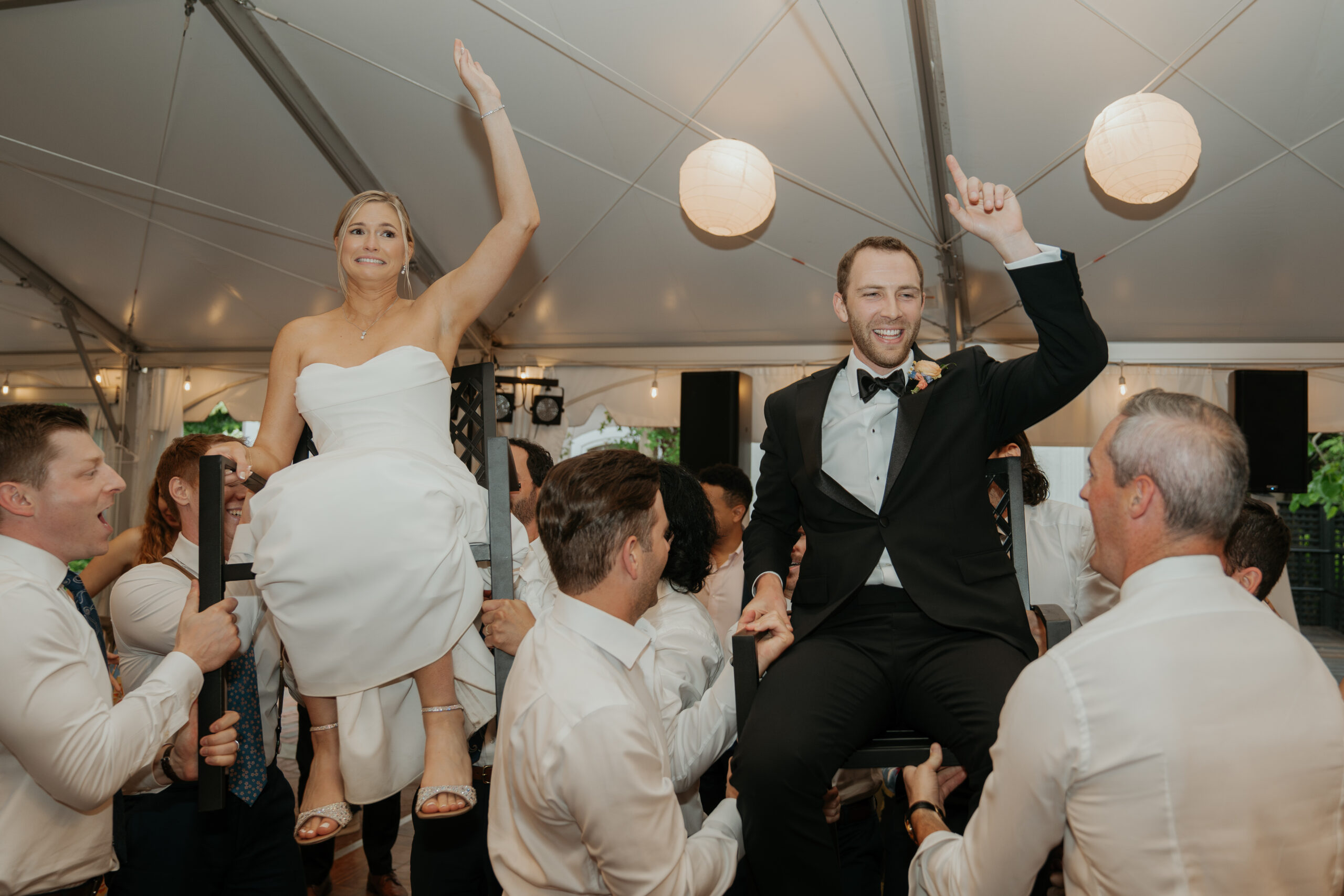 Bride and groom celebrating at their summer garden party wedding reception doing the Hora. The bride looks stressed while the groom looks like hes having fun. a bunch of guests are lifting them up by their chairs.