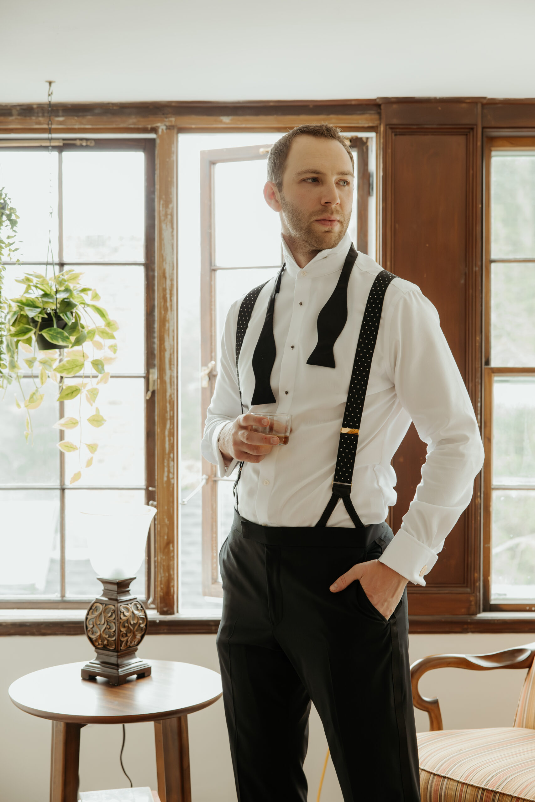 Groom standing in his suit pants, suspenders, dress shirt with his bowtie untied. He's holding a glass of whiskey in one hand while his other hand is in his pocket, and he is looking off in the distance. He is getting ready for his summer garden party wedding at Long Hill in Beverly, MA.
